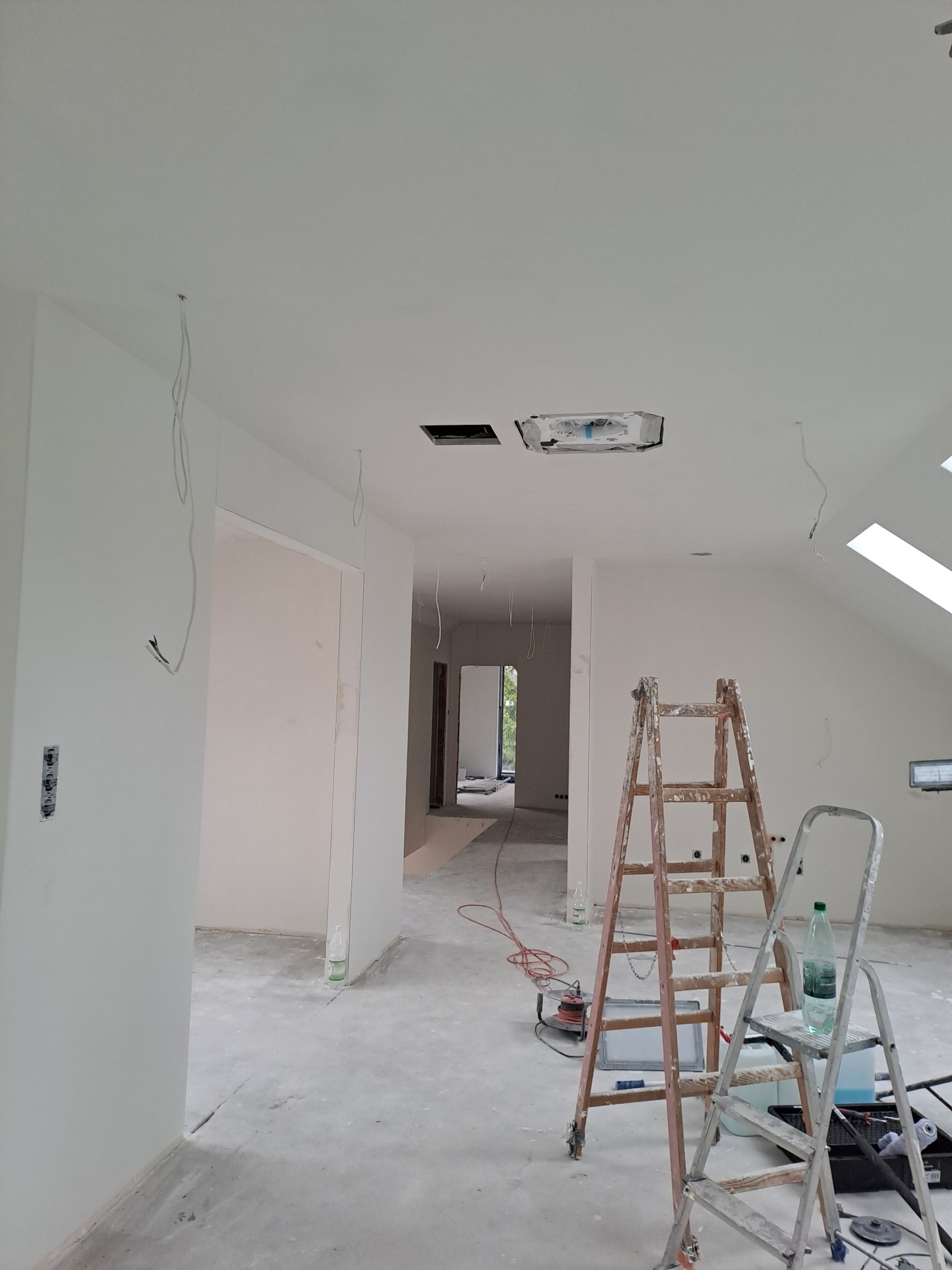 Jasne wnętrze w trakcie prac wykończeniowych: białe ściany, dwie drabiny malarskie, widoczne przewody elektryczne i narzędzia. W tle okno dachowe i otwarte przejście.