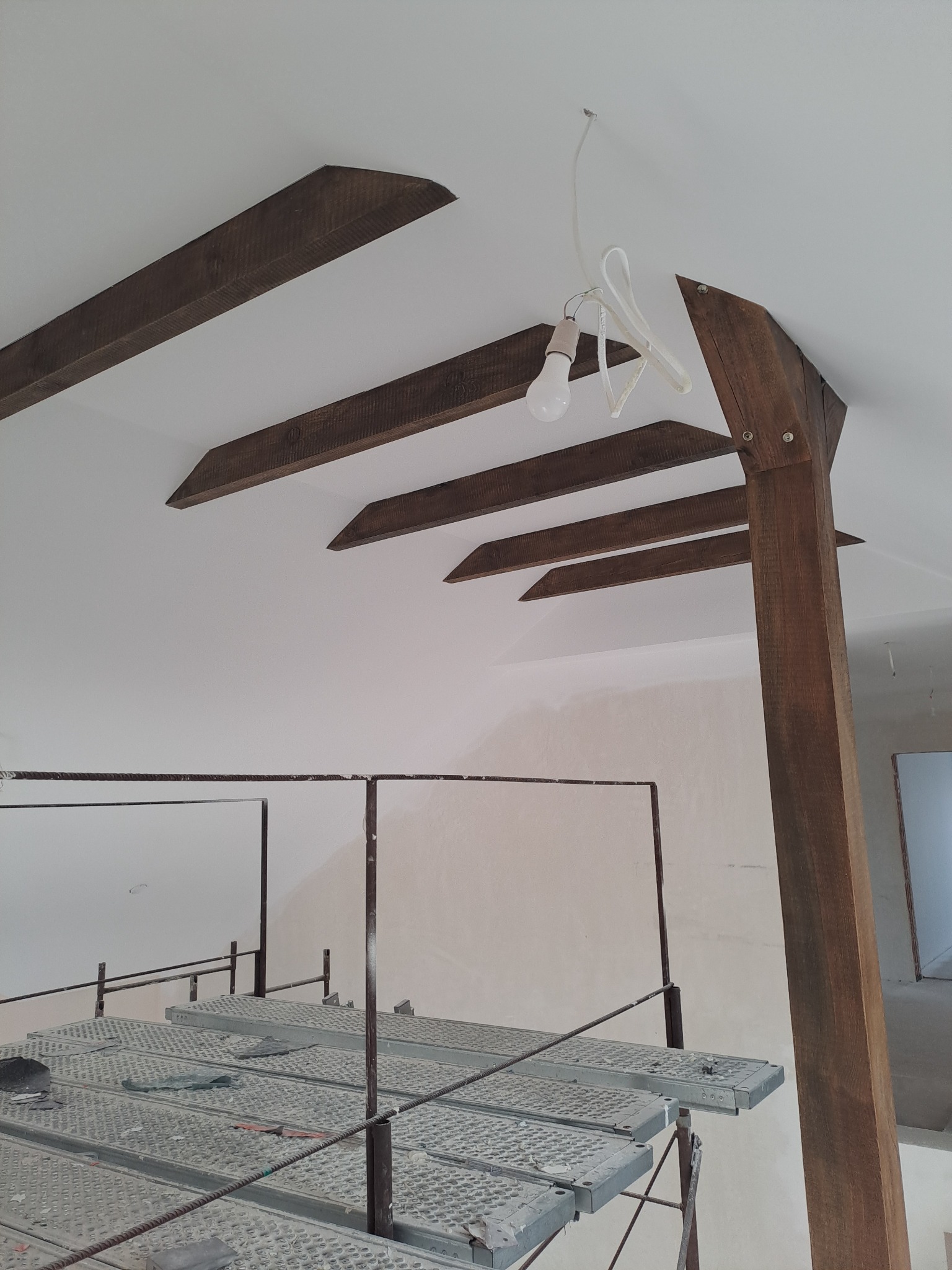 Białe wnętrze z drewnianymi belkami stropowymi, widoczne rusztowanie i wisząca żarówka. W tle ściana w jasnym odcieniu beżu.