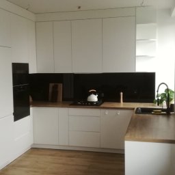 Firma Handlowo Usługowa ADIKO Adrian Zygmuntowicz - Biała kuchnia na wymiar z drewnianym blatem, czarnym panelem i zlewozmywakiem. Minimalistyczny design z czajnikiem na płycie i rośliną w tle.