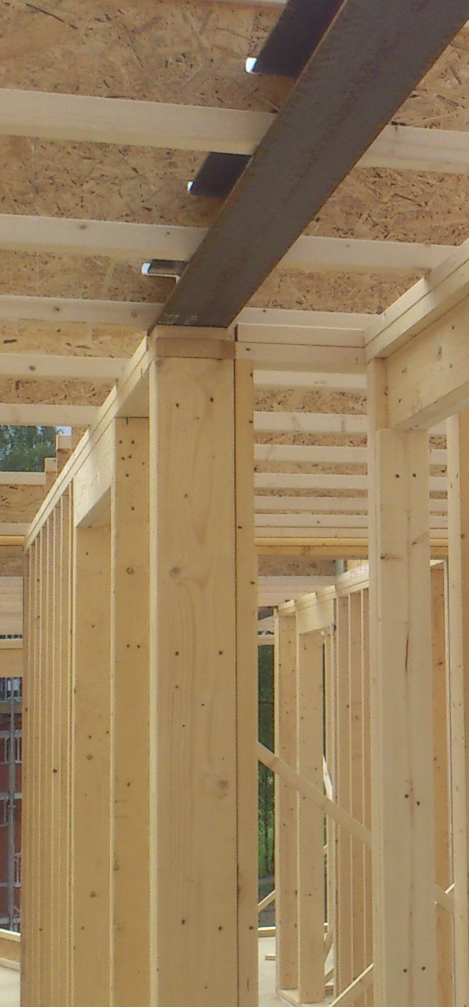 Konstrukcja szkieletowa z drewna i metalu w trakcie budowy. Widoczne elementy ścian, stropu i metalowe wzmocnienie. Jasne drewno, surowe wykończenie.