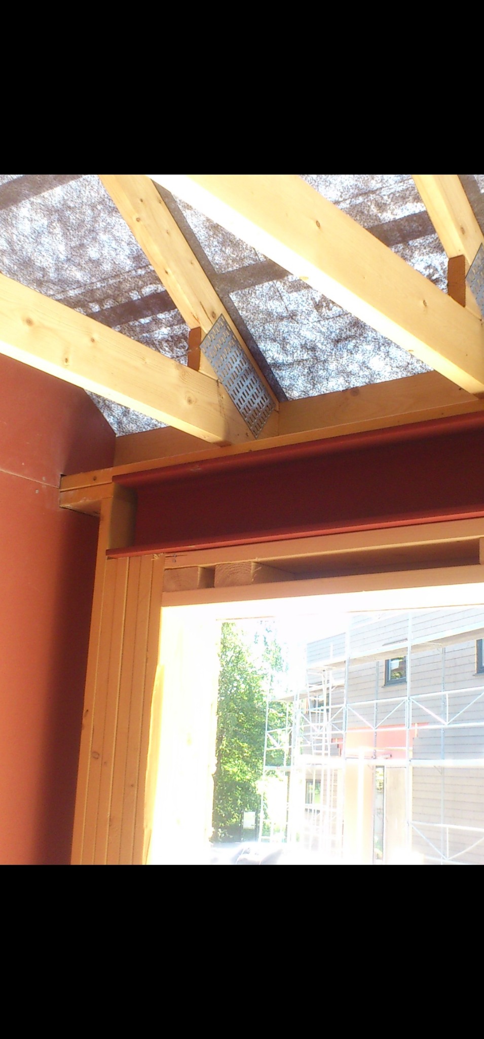 Konstrukcja dachu z drewnianymi belkami, połączonymi metalowymi płytkami, widok z wnętrza budynku w trakcie budowy. Czerwona belka stalowa nad otworem okiennym.