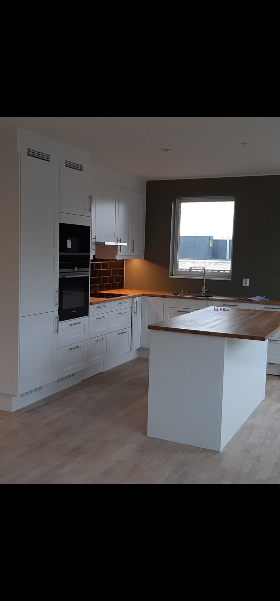 Jasna kuchnia z drewnianym blatem i wyspą kuchenną, brązowe płytki nad blatem, okno z widokiem na budynek przemysłowy, minimalistyczny design.