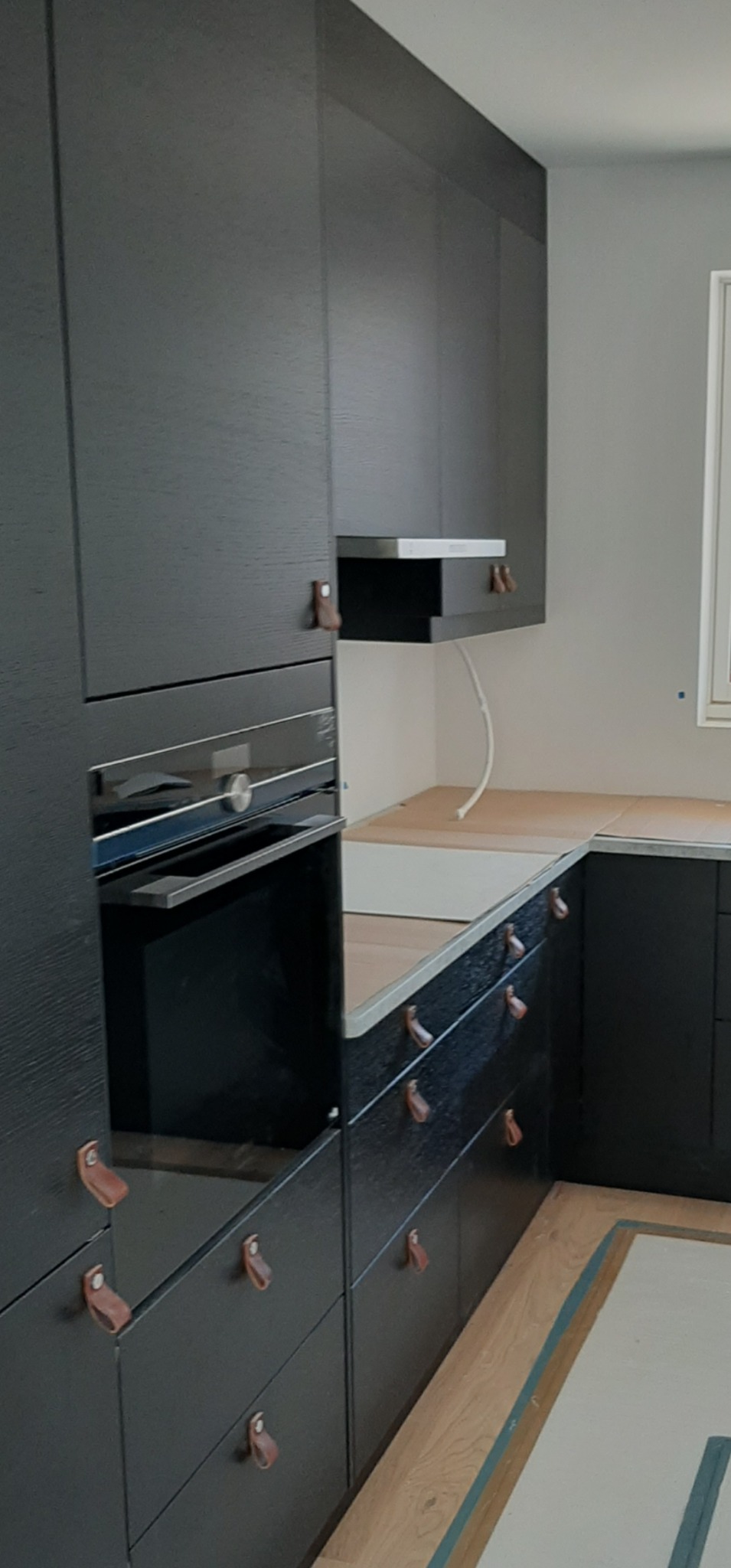 Ciemna zabudowa kuchenna z piekarnikiem i szafkami z nietypowymi, skórzanymi uchwytami. Częściowo widoczny blat i podłoga.