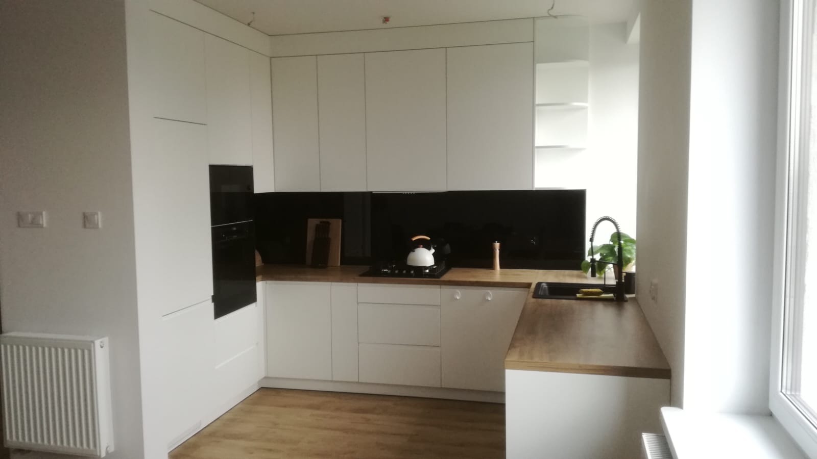 Biała kuchnia na wymiar z drewnianym blatem, czarnym panelem i zlewozmywakiem. Minimalistyczny design z czajnikiem na płycie i rośliną w tle.