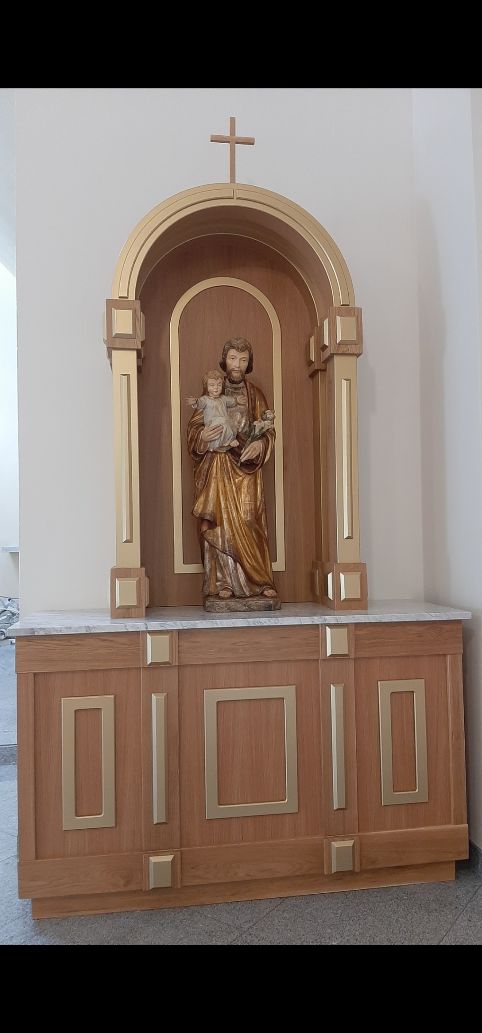 Drewniana szafka z marmurowym blatem i niszą zwieńczoną krzyżem, w której stoi figura św. Józefa z Dzieciątkiem. Dekoracyjne złote detale na frontach szafki.