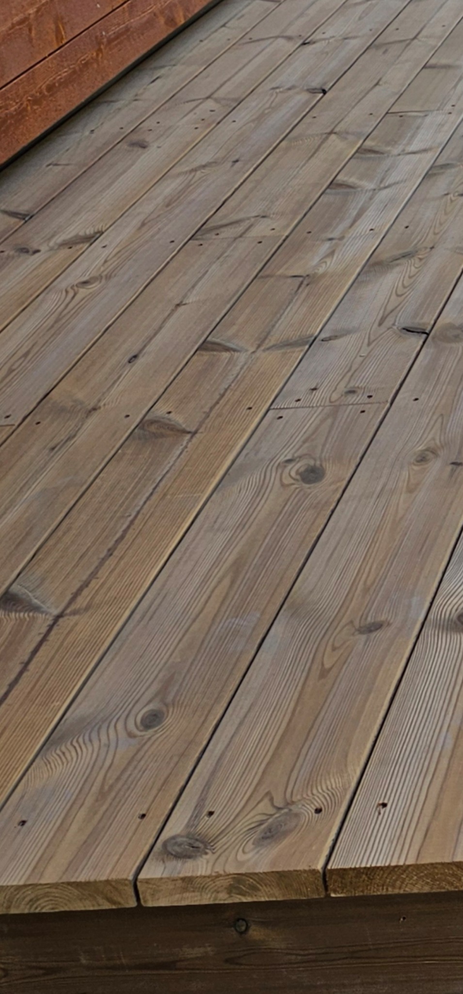 Ukończony taras z desek z widocznymi słojami drewna i gwoździami, ułożony pod kątem, z fragmentem brązowej ściany w tle. Drewno ma naturalny, ciepły odcień.