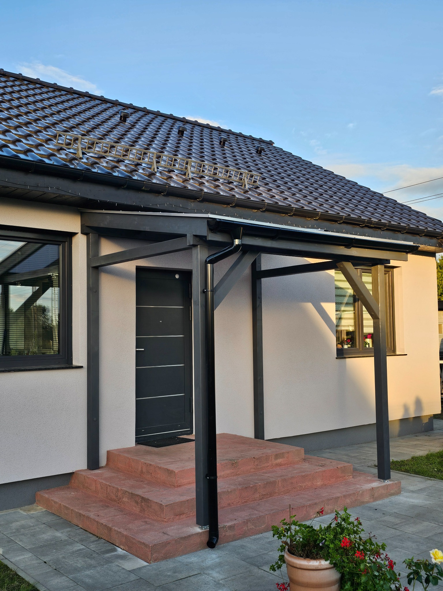 Nowoczesny dom z zadaszeniem nad wejściem, wspartym na ciemnych, drewnianych słupach. Czerwone schody prowadzą do ciemnych drzwi. Uwagę przykuwa minimalistyczny design.