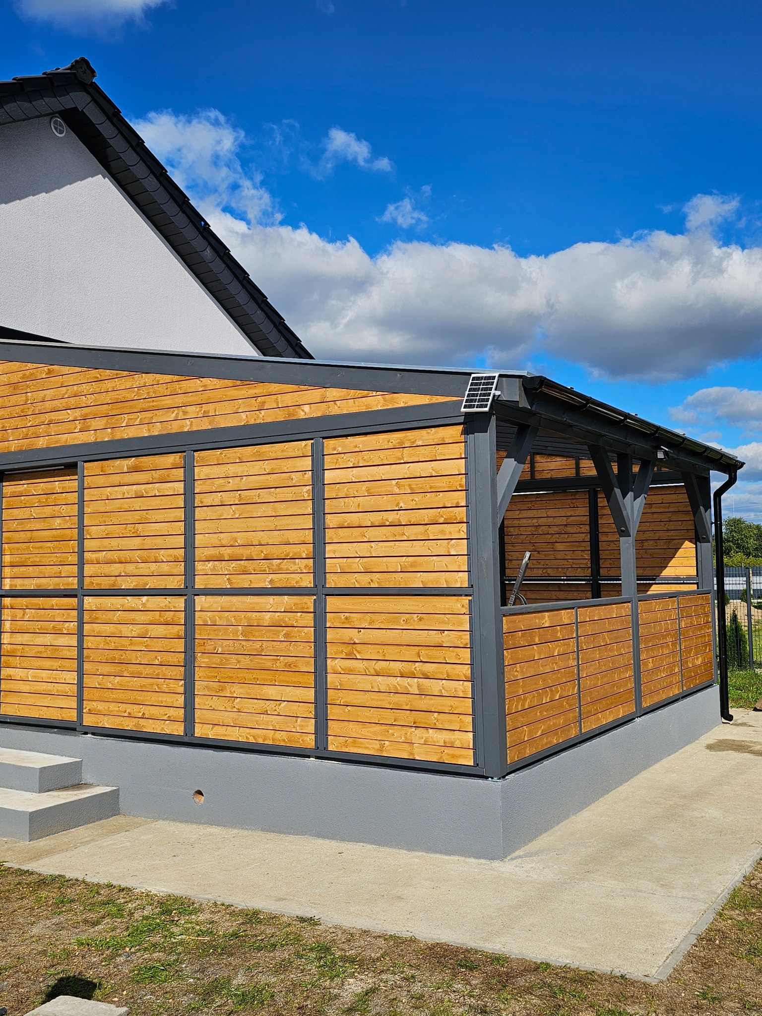 Nowoczesna, drewniana pergola z szarymi elementami konstrukcyjnymi i panelem solarnym na dachu, na tle błękitnego nieba z chmurami. Widok z zewnątrz, z boku.