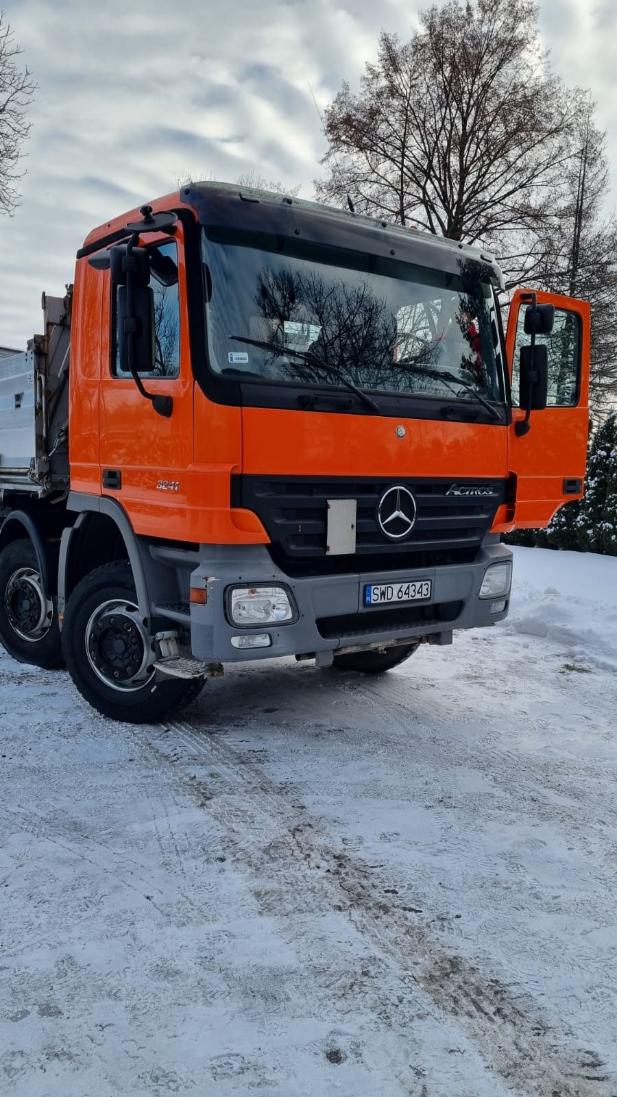 Pomarańczowy samochód ciężarowy Mercedes Actros z otwartymi drzwiami, zaparkowany na śniegu. Widoczne drzewa w tle i tablica rejestracyjna.