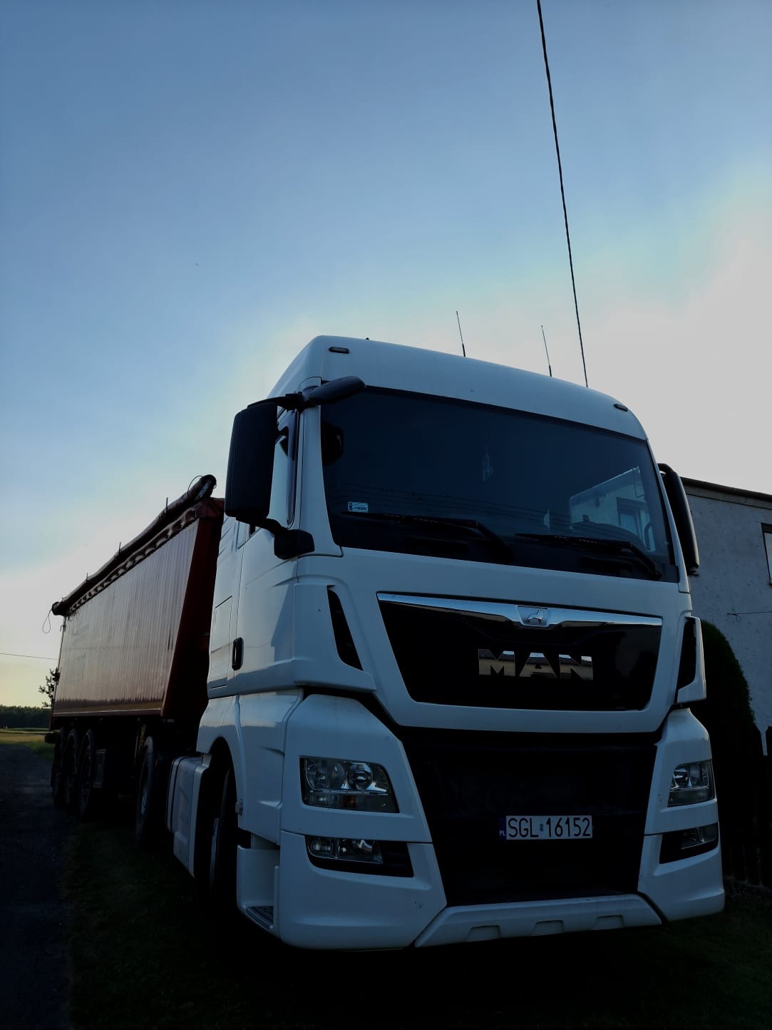 Biały samochód ciężarowy MAN z czerwoną naczepą, zaparkowany na trawie, z numerem rejestracyjnym SGL 16152, sfotografowany pod kątem z dołu w pochmurny dzień.