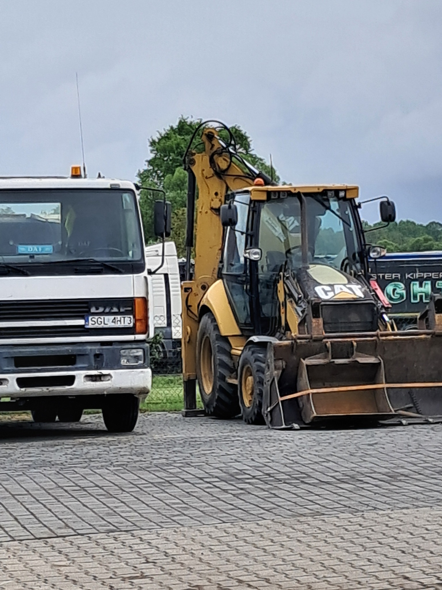 Żółta koparko-ładowarka CAT i biała ciężarówka DAF na kostce brukowej. Widoczne tablice rejestracyjne. Tło rozmyte, zieleń drzew. Pochmurne niebo.