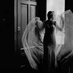 Czarno-białe zdjęcie kobiety w długiej sukni, stojącej przy otwartych drzwiach z powiewającą tkaniną, tworzącej efektowną stylizację w klimacie vintage.