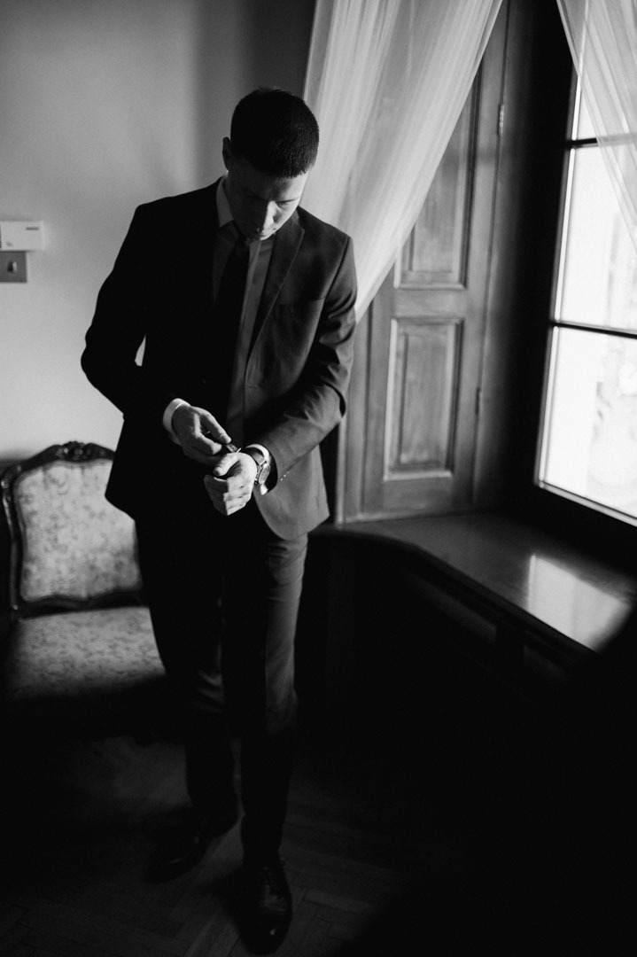 Czarno-białe zdjęcie mężczyzny w garniturze zapinającego zegarek przy oknie z firanką. Ujęcie wewnątrz pomieszczenia, widoczny fotel w stylu retro.