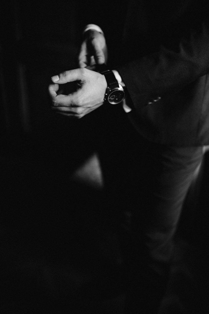 Czarno-białe zbliżenie na męską dłoń zapinającą zegarek na tle ciemnego garnituru. Skupienie na detalu i elegancji, podkreślone przez minimalistyczną estetykę.