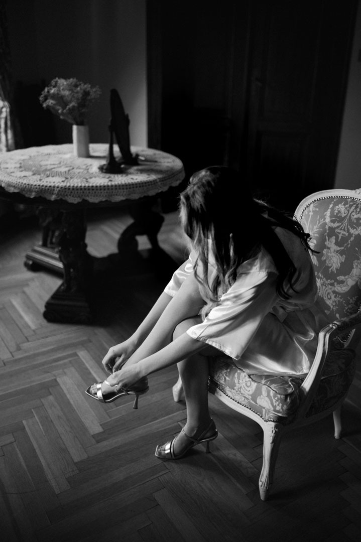 Czarno-białe zdjęcie kobiety w satynowym szlafroku, siedzącej na zdobionym krześle i zapinającej srebrne buty na obcasie. W tle okrągły stolik z kwiatami.