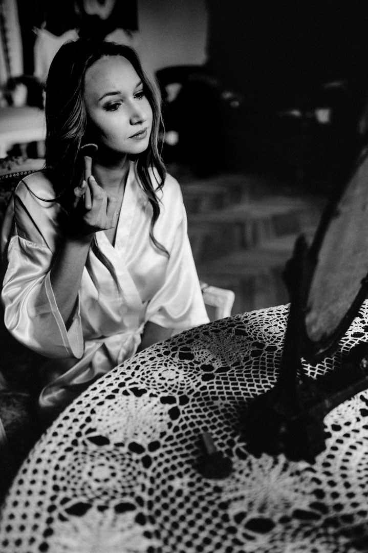 Kobieta w szlafroku, w czerni i bieli, nakłada makijaż przed lustrem na stole z koronkowym obrusem. Subtelne oświetlenie podkreśla detale.