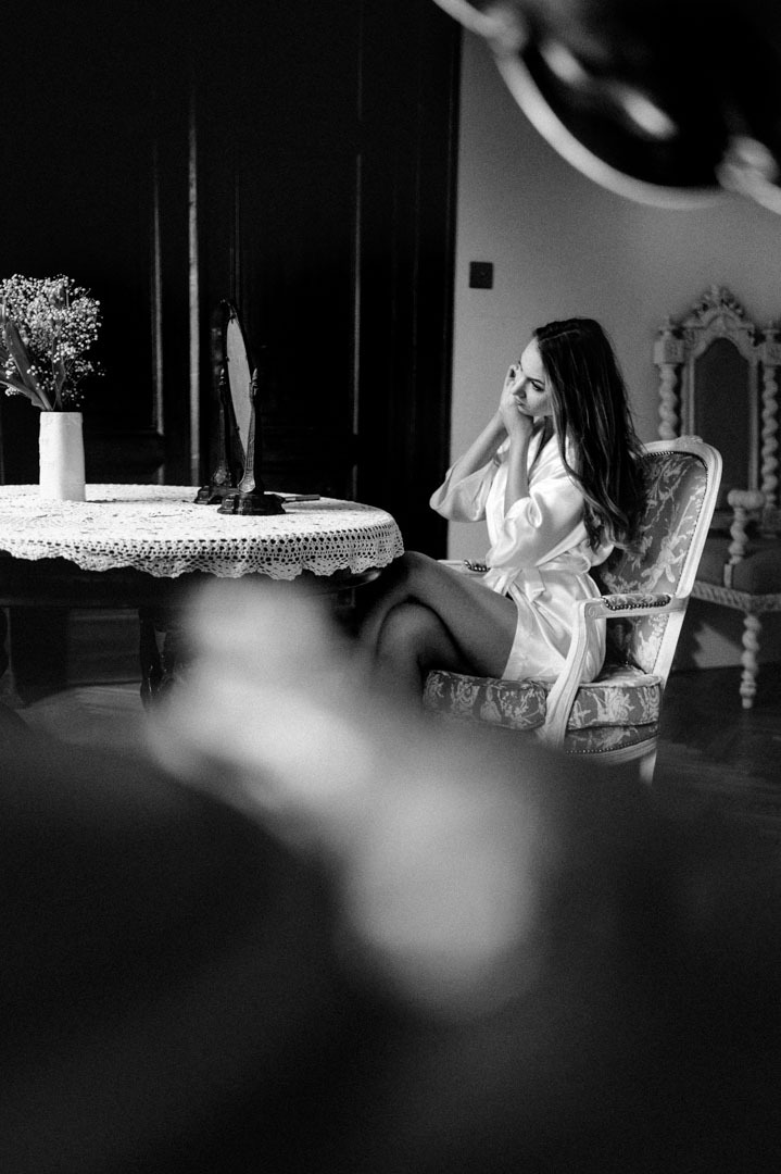 Czarno-białe zdjęcie kobiety w szlafroku, siedzącej przy toaletce z lustrem, przygotowującej się. W tle ciemne drzwi i wazon z kwiatami. Ujęcie zza zasłony.