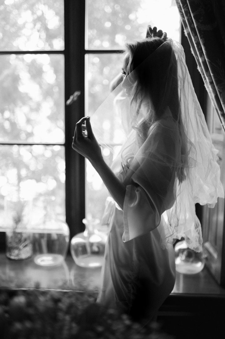 Czarno-białe zdjęcie panny młodej w szlafroku, stojącej przy oknie z welonem w dłoni. Na parapecie dekoracyjne flakony, za oknem zieleń.
