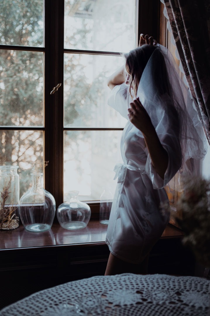 Panna młoda w białym szlafroku i welonie spogląda przez okno, obok szklane wazony z suszem na drewnianym parapecie. Ujęcie w naturalnym świetle.
