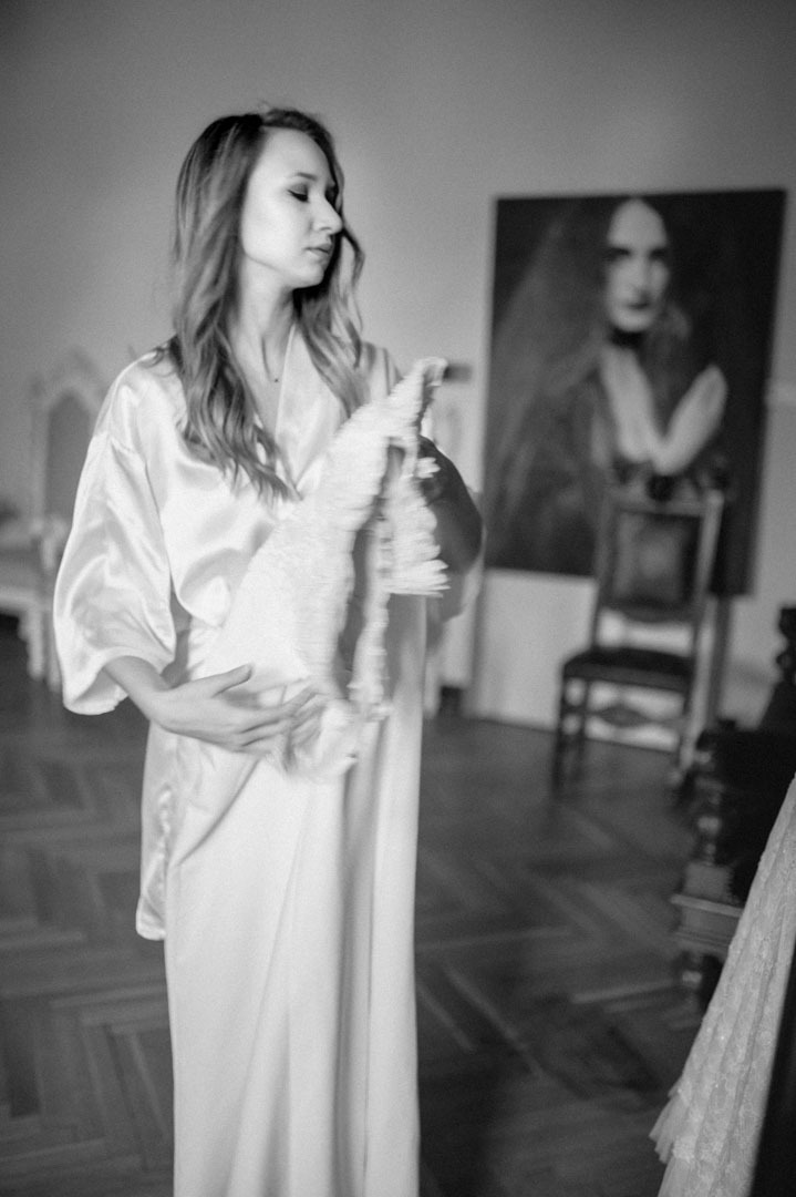 Czarno-białe zdjęcie panny młodej w szlafroku, trzymającej suknię ślubną. W tle obraz na ścianie i krzesło. Parkiet na podłodze.