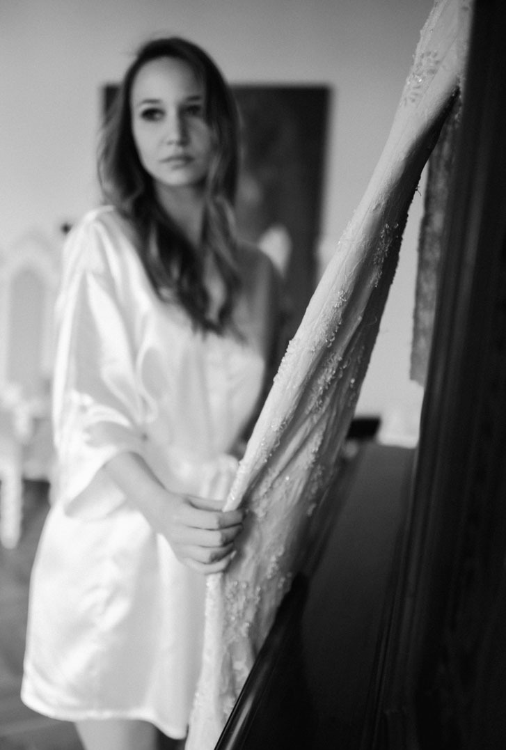 Panna młoda w szlafroku trzyma suknię ślubną z delikatnymi zdobieniami, uchwycona w czerni i bieli w Wieluniu. Subtelne przygotowania.