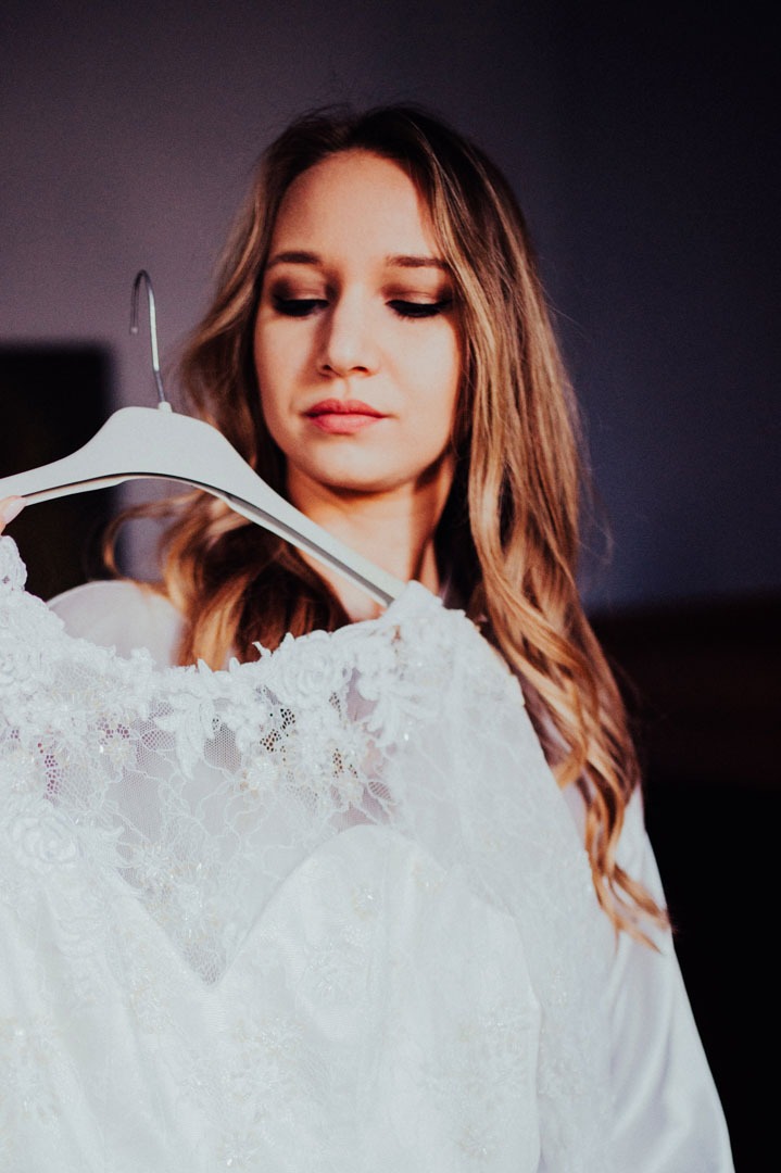 Panna młoda z blond włosami spogląda na białą, koronkową suknię ślubną wiszącą na wieszaku, w tle ściana w odcieniach fioletu. Romantyczne oświetlenie.