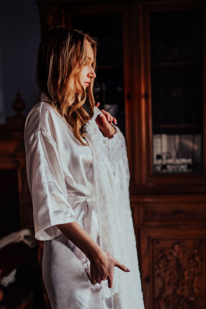 Panna młoda w satynowym szlafroku trzymająca suknię ślubną na tle ciemnego, zdobionego mebla. Profil, ciepłe światło.