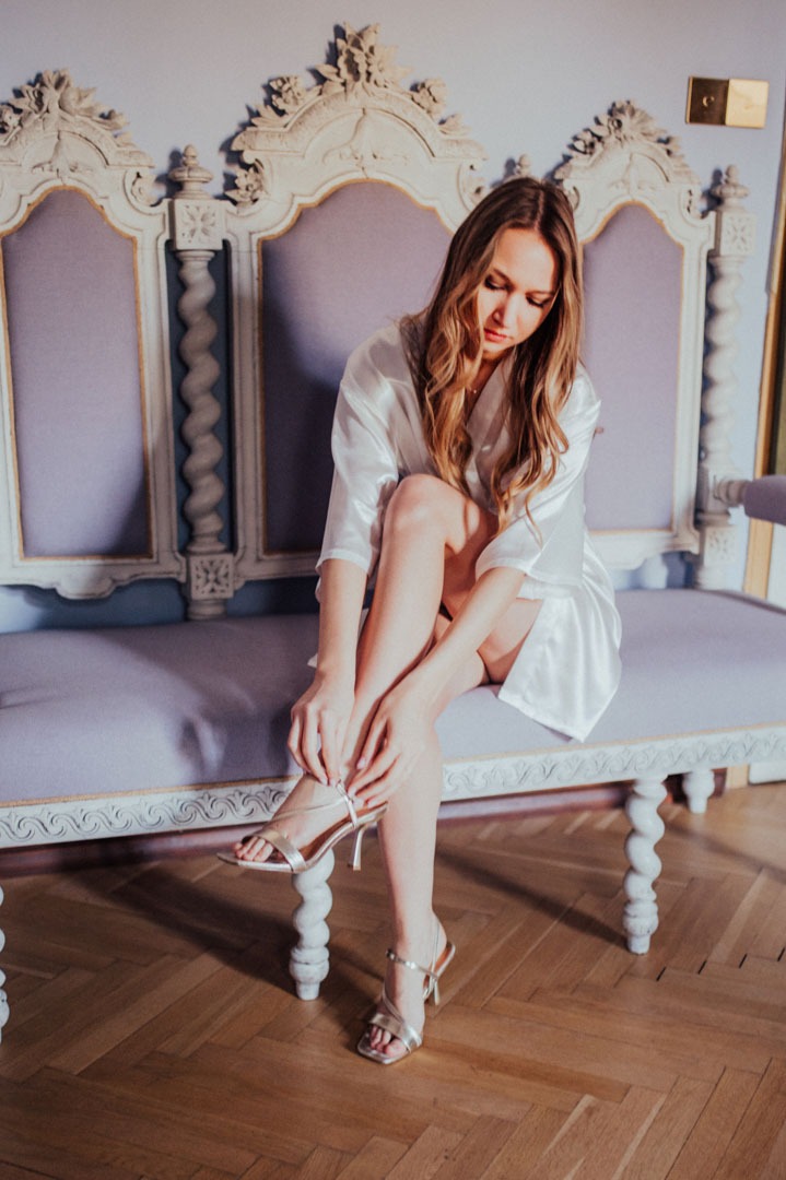 Kobieta w satynowym szlafroku, siedząca na zdobionej ławce, zapinająca złote sandały na wysokim obcasie na tle błękitnej ściany i drewnianej podłogi.