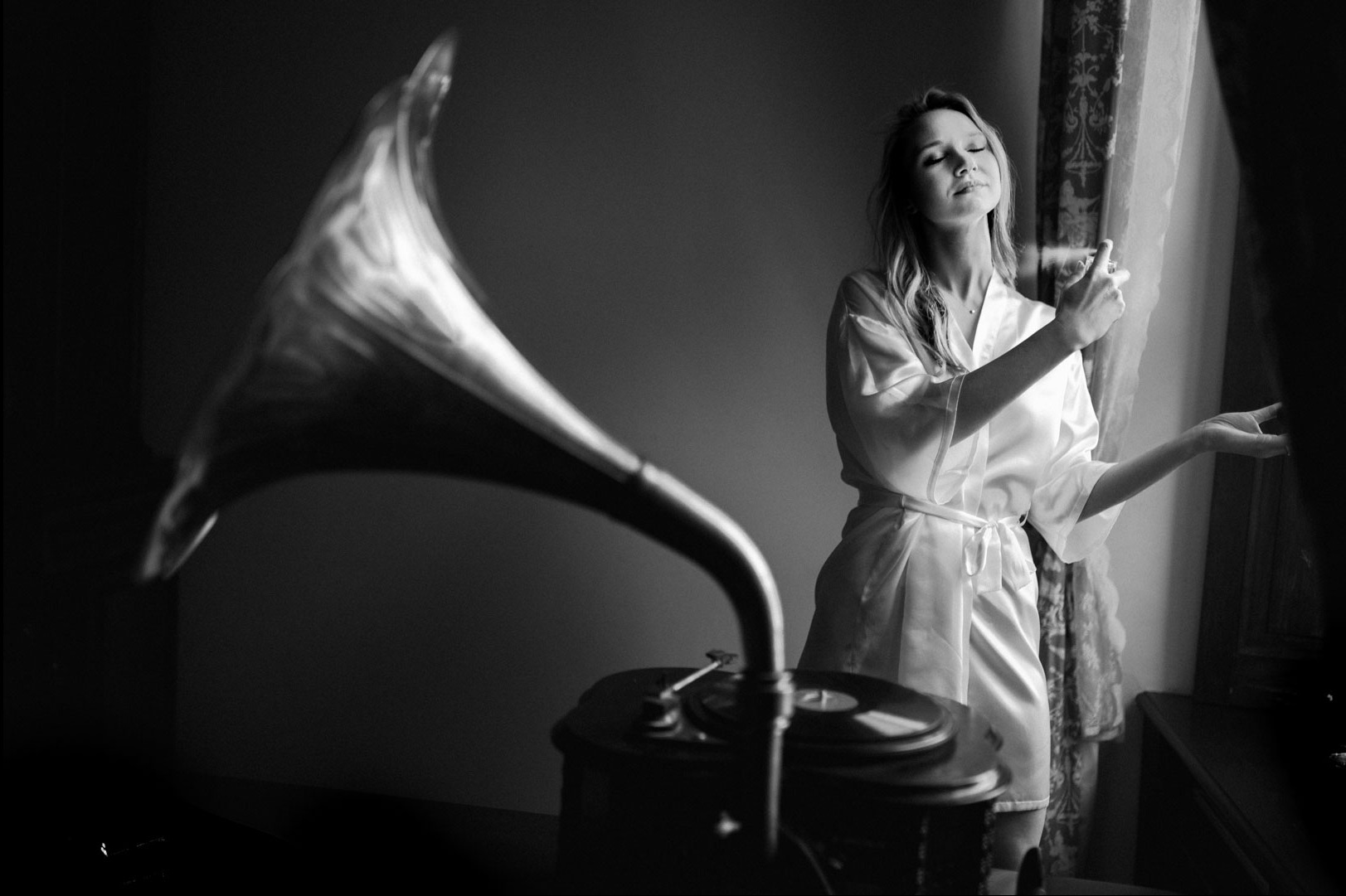 Czarno-białe zdjęcie kobiety w szlafroku, stojącej przy oknie i spryskującej się perfumami. W tle gramofon z tubą. Klimatyczne ujęcie.