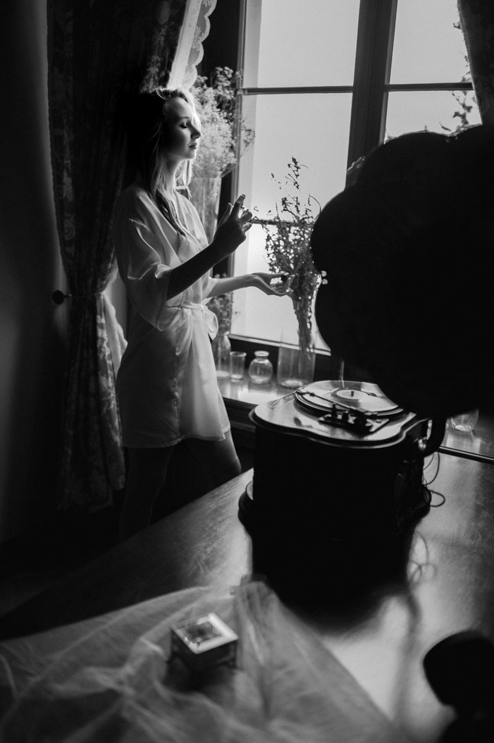 Czarno-białe zdjęcie kobiety w szlafroku, spryskującej się perfumami przy oknie, obok gramofonu. U dołu pudełko na biżuterię i fragment tiulu. Delikatne, naturalne światło.