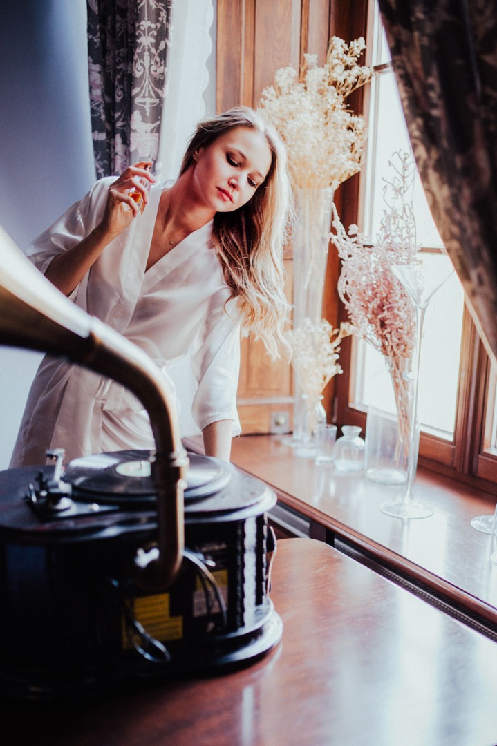 Kobieta w szlafroku, z flakonem perfum, pozuje obok gramofonu na tle okna z bukietami w wazonach. Ciepłe, naturalne światło.