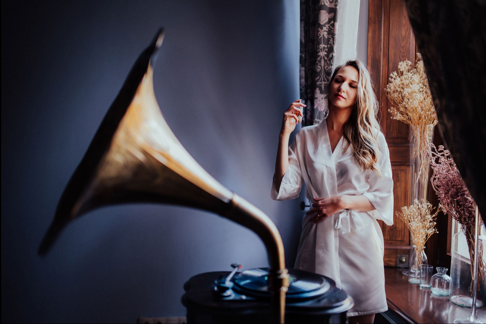 Kobieta w szlafroku pozuje obok gramofonu na korbkę z tubą, w tle okno z zasłonami i suszone kwiaty w wazonach. Subtelne światło i stonowana kolorystyka.