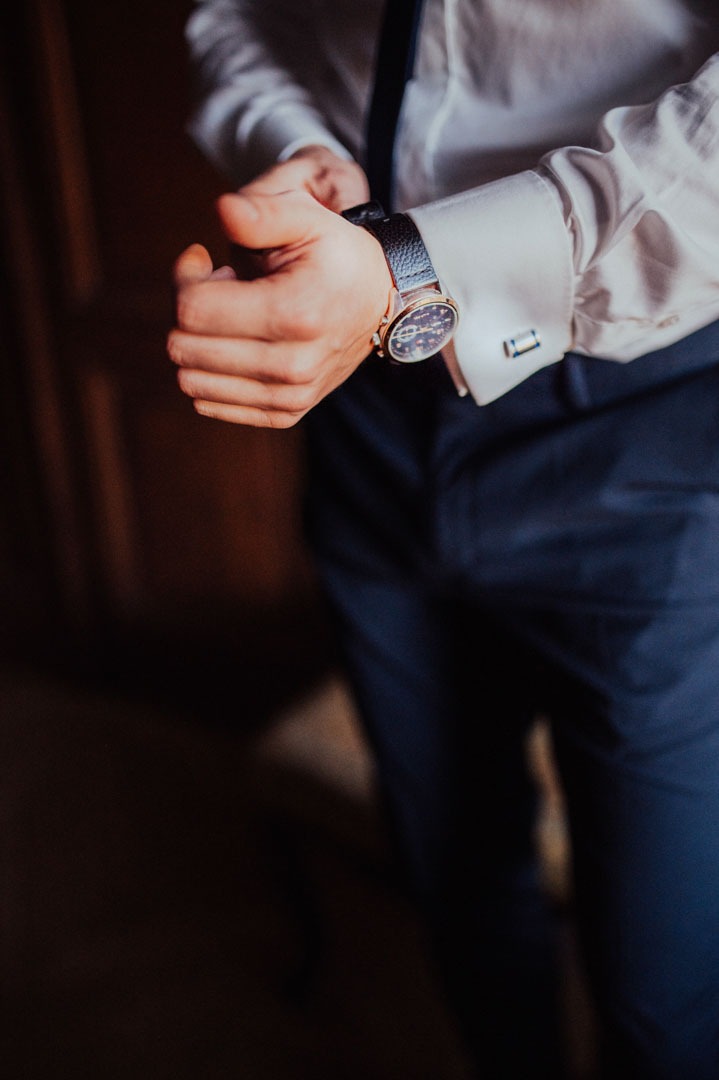 Elegancki mężczyzna w białej koszuli i granatowych spodniach zapina zegarek z czarnym paskiem i mankietami na tle ciemnego drewna.