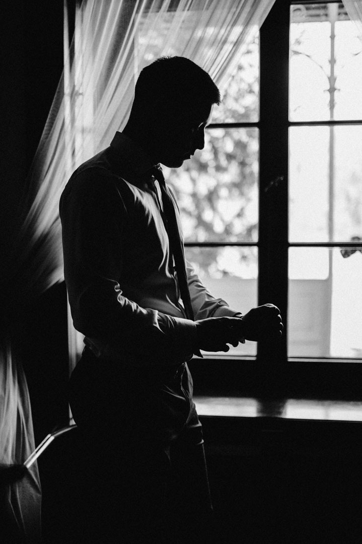 Czarno-białe zdjęcie sylwetki mężczyzny w koszuli i krawacie, stojącego przy oknie z firankami, zapinającego mankiet koszuli. Silny kontrast światła i cienia.