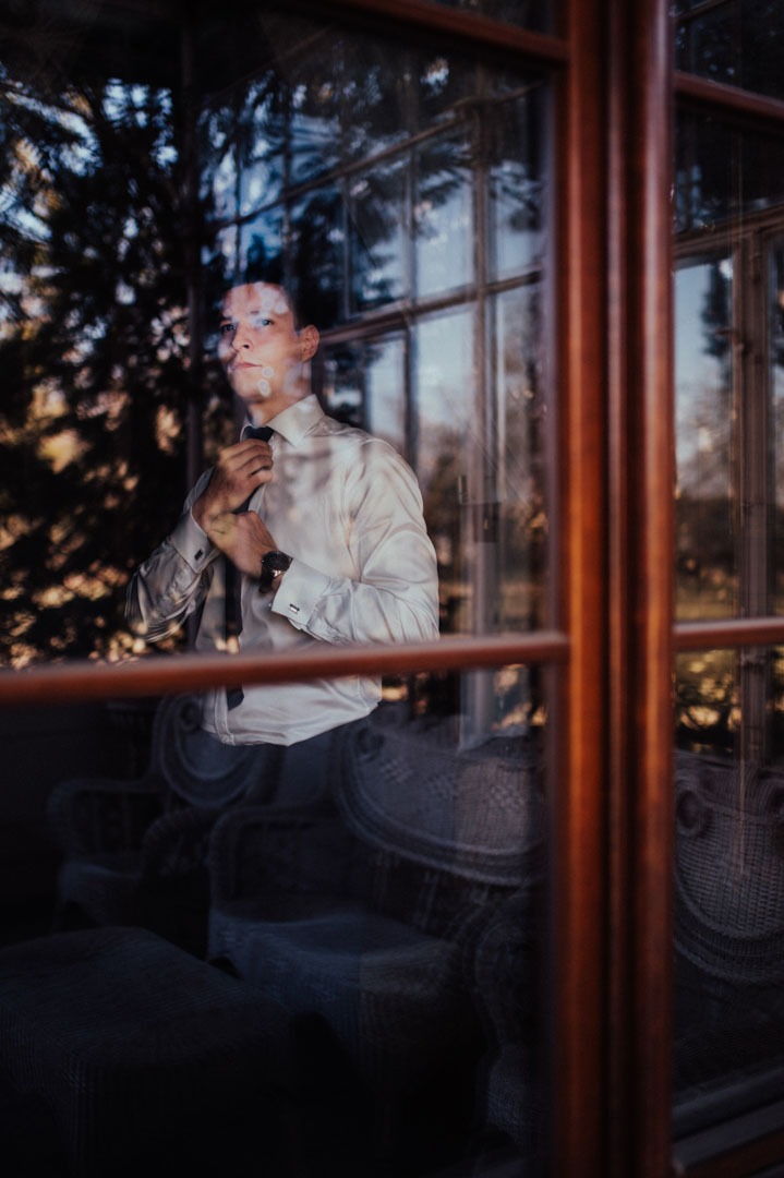 Elegancki mężczyzna w białej koszuli zawiązuje krawat, widoczny przez okno z drewnianą ramą. Na pierwszym planie odbicie wnętrza z kanapą.