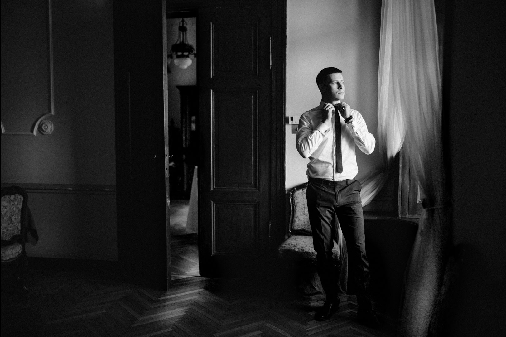 Elegancki mężczyzna w białej koszuli i krawacie, stojący w pokoju z otwartymi drzwiami i firankami, w czerni i bieli, zapinający krawat.