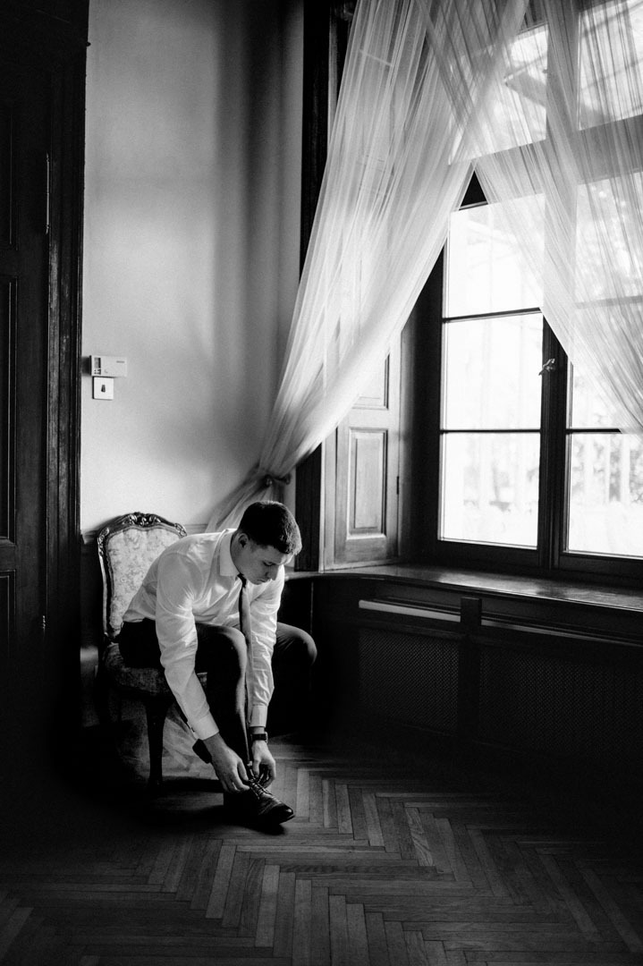 Czarno-białe zdjęcie mężczyzny w koszuli i krawacie, siedzącego na krześle przy oknie z firankami, zawiązującego sznurówki butów. Parkiet na podłodze.