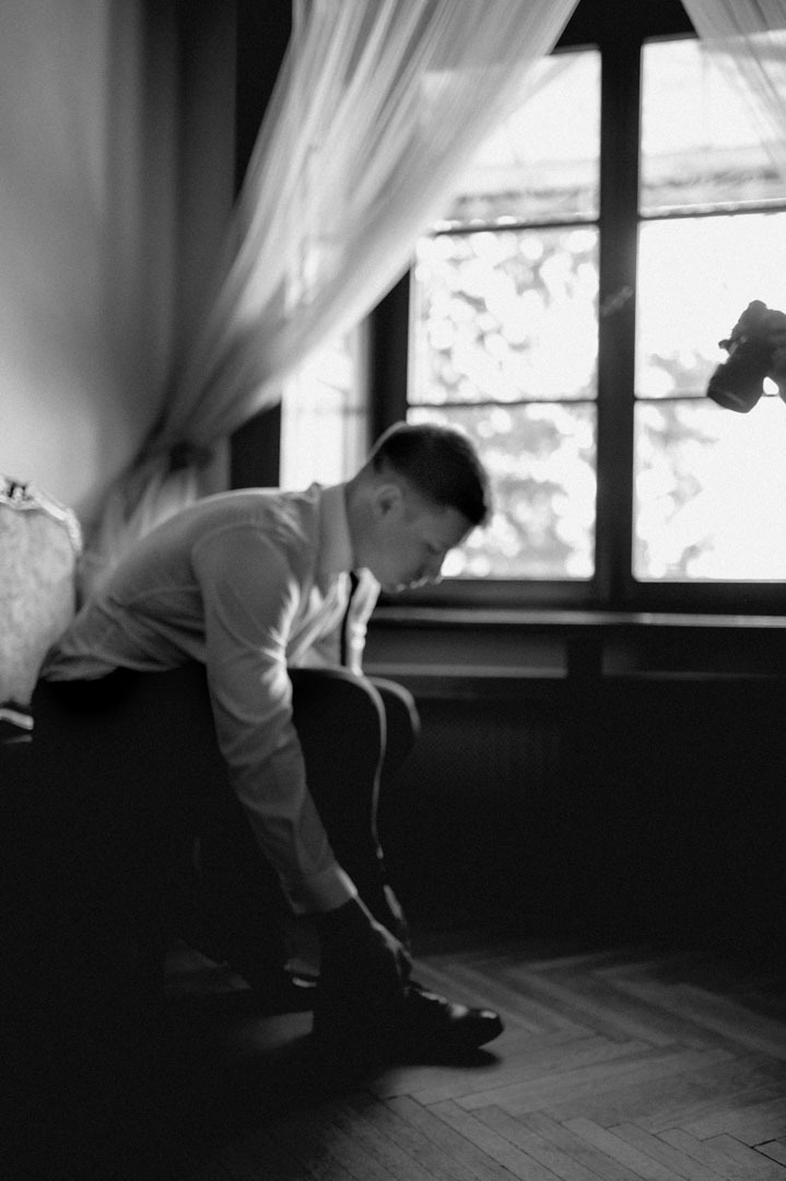 Czarno-białe zdjęcie mężczyzny w koszuli, siedzącego na kanapie i sznurującego buty przy oknie. W tle widoczny fragment aparatu fotografa.