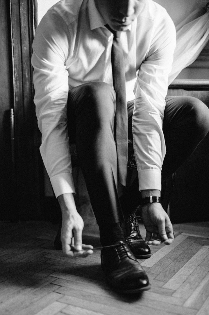 Czarno-białe zdjęcie mężczyzny w koszuli i krawacie, klęczącego i wiążącego sznurówki eleganckich butów na drewnianej podłodze, z ciemnymi drzwiami w tle.