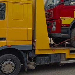 Pomoc Drogowa Lublin - Piotr Wiernio - Żółta laweta przewozi czerwony wóz strażacki. Widoczne zabezpieczenia łańcuchami. Ujęcie z boku, na pierwszym planie koła lawety i fragment budynku w tle.