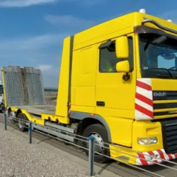 Ż&oacute;łta laweta DAF XF na autostradzie z zaparkowanym białym tirem SCANIA. Widoczny fragment barierek i krajobrazu. Słoneczny dzień.