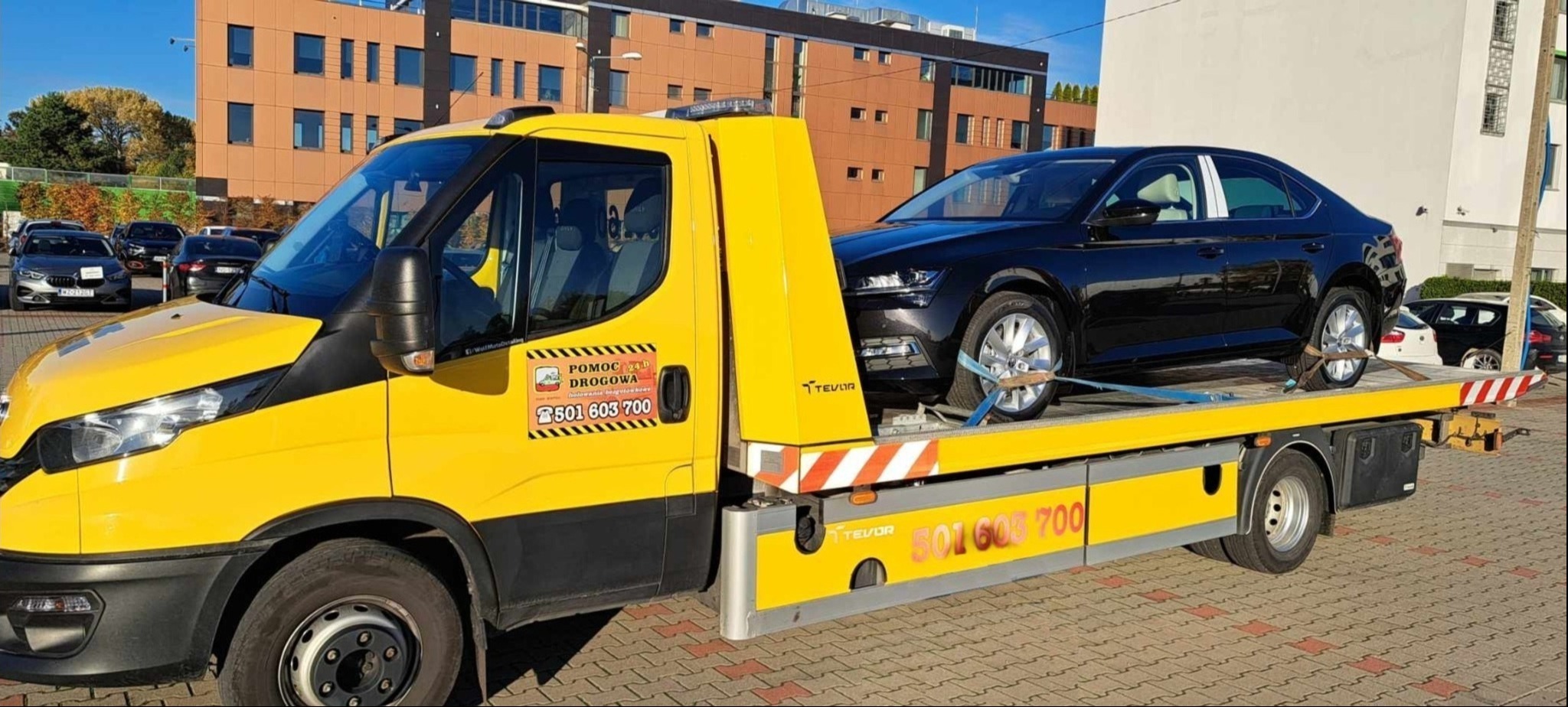 Żółta laweta z czarnym samochodem osobowym na platformie, zabezpieczonym pasami. Na lawecie widoczne logo firmy i numer telefonu. Budynek w tle.
