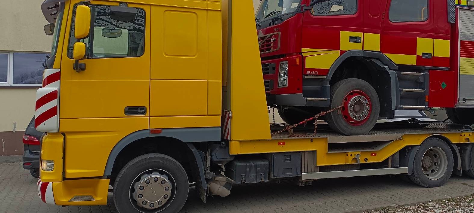 Żółta laweta przewozi czerwony wóz strażacki. Widoczne zabezpieczenia łańcuchami. Ujęcie z boku, na pierwszym planie koła lawety i fragment budynku w tle.