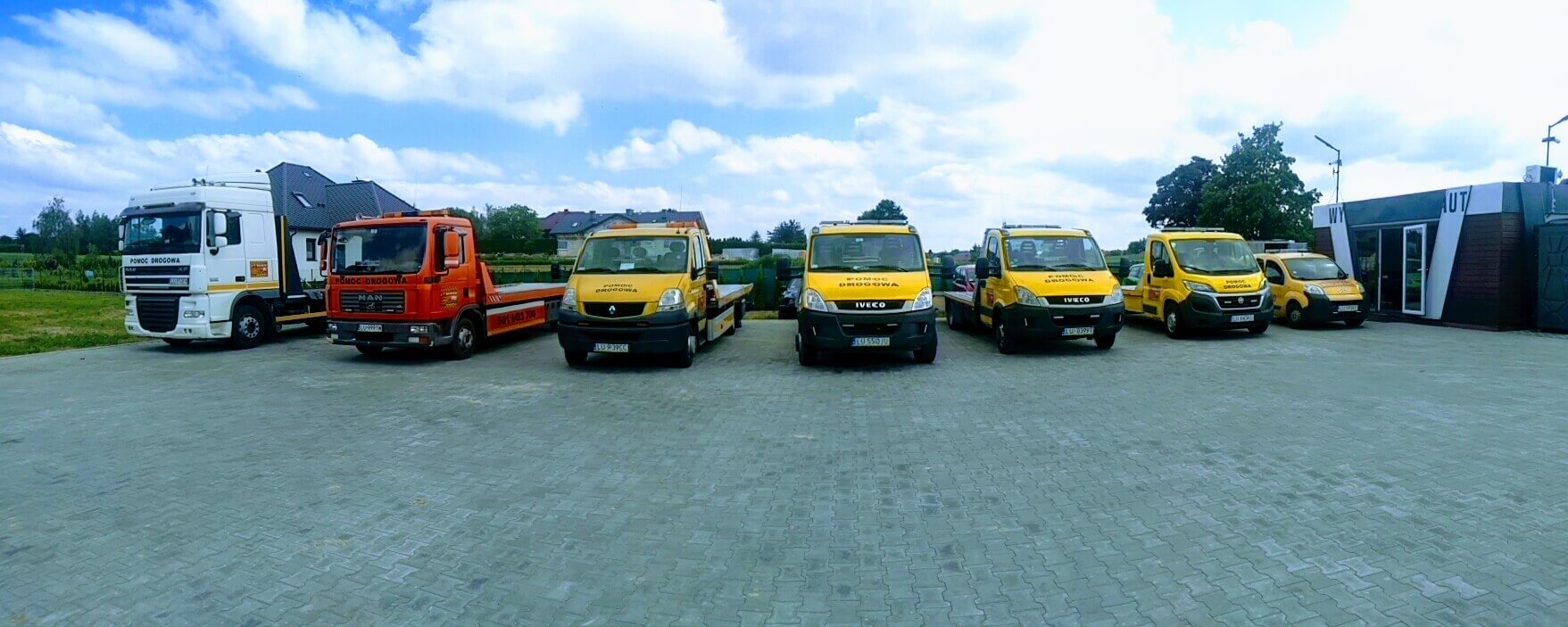 Flota pojazdów pomocy drogowej, w tym lawety MAN i Iveco, ustawiona rzędem na parkingu przed budynkiem biurowym w słoneczny dzień. Widoczne logo firmy na pojazdach.