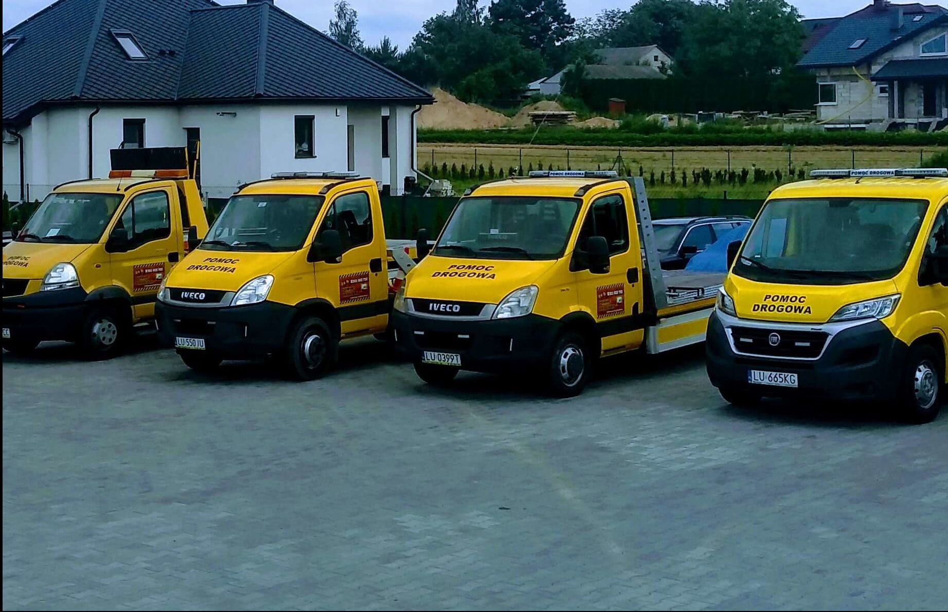 Żółte lawety Iveco i Fiat z napisem 'Pomoc Drogowa' zaparkowane na szarym bruku przed budynkiem. Widoczne tablice rejestracyjne i elementy konstrukcyjne lawet.