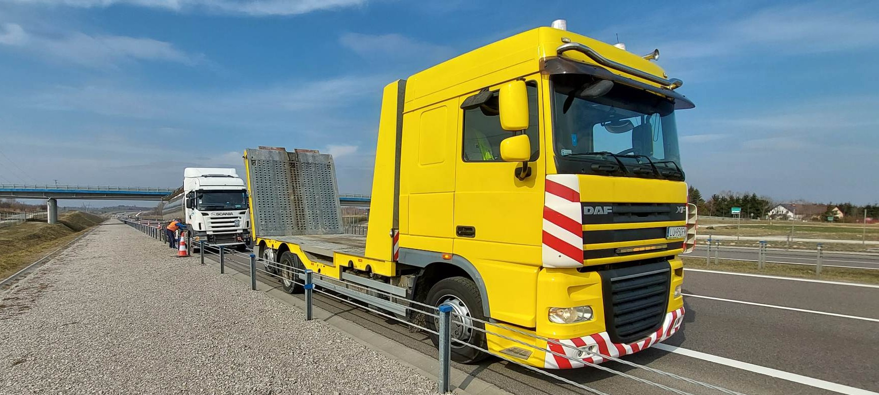 Żółta laweta DAF XF na autostradzie z zaparkowanym białym tirem SCANIA. Widoczny fragment barierek i krajobrazu. Słoneczny dzień.