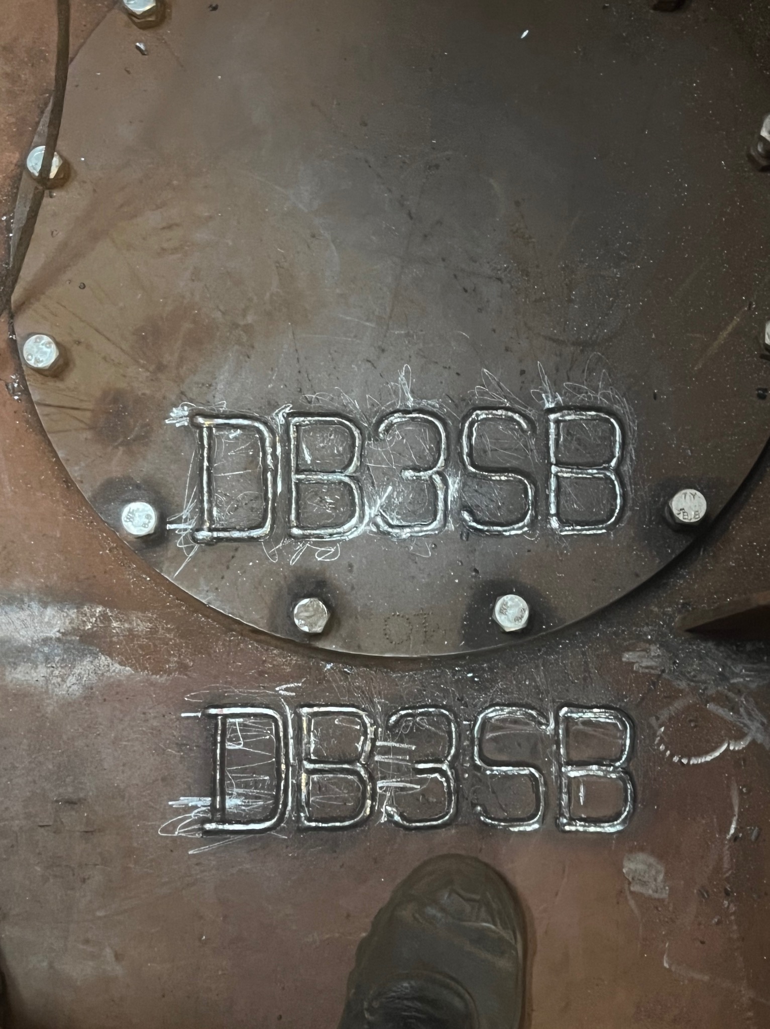 Metalowa płyta z wyciętym napisem DB3SB, widoczne ślady obróbki i spawania, całość przymocowana śrubami. W tle fragment buta.