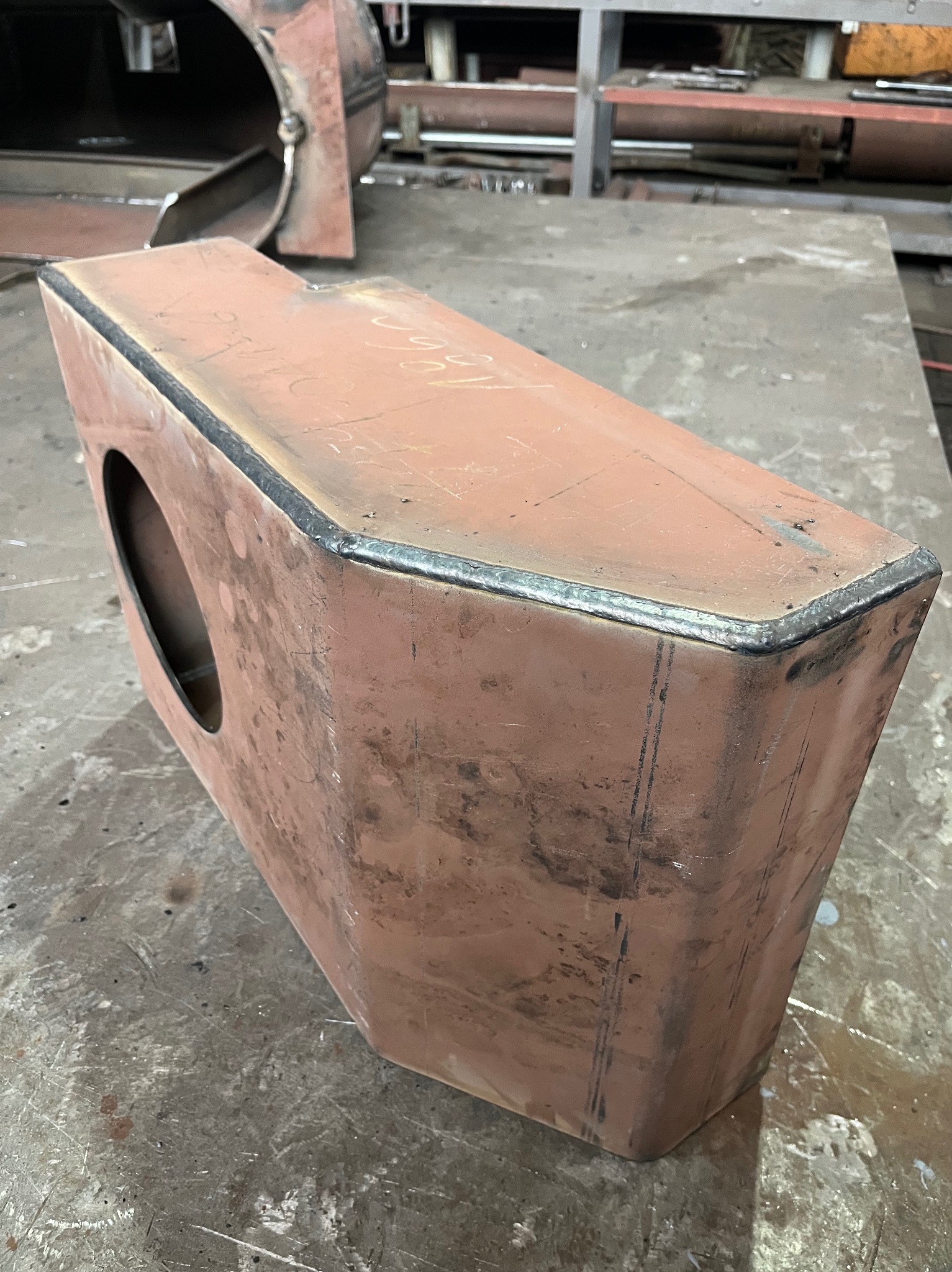 Spawana konstrukcja skrzynkowa z otworem, wykonana z metalu w kolorze rdzawym, z widocznymi spawami na krawędziach, leżąca na warsztatowym stole.