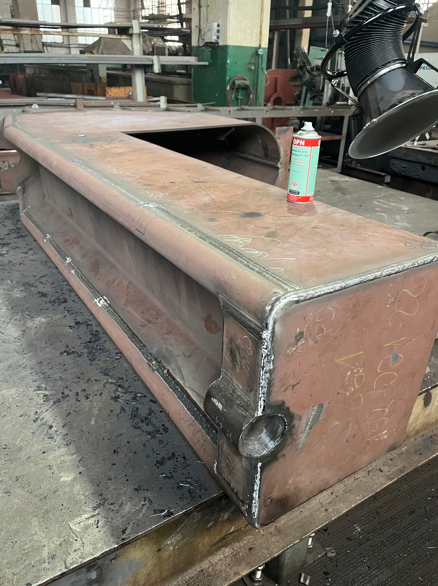 Metalowa konstrukcja po obróbce, widoczne spawy, szlifowanie i ślady po cięciu. Na górze puszka ze sprayem. W tle warsztat.