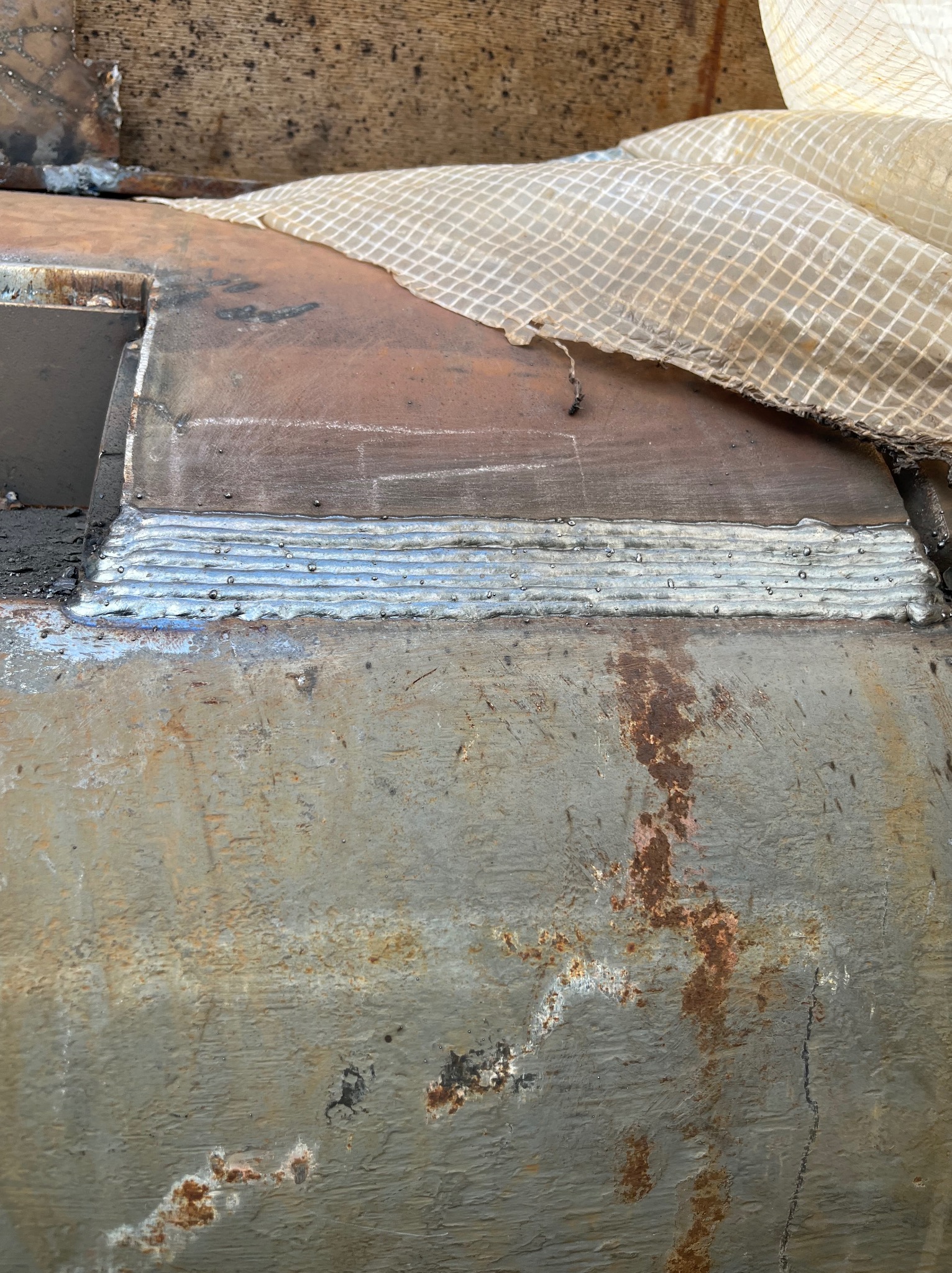 Metalowa powierzchnia z widocznymi śladami spawania w postaci równych, srebrnych linii. Fragment konstrukcji z rdzawymi plamami i resztkami farby. Częściowo przykryta tkaniną.